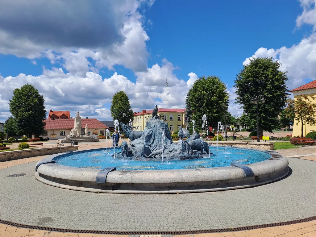 Fontanna z rzeźbami przedstawiającymi postaci ludzkie otoczona tryskającą wodą, wokół widoczne zadbane drzewa, budynki oraz fragment brukowanego placu miejskiego pod błękitnym niebem z chmurami.