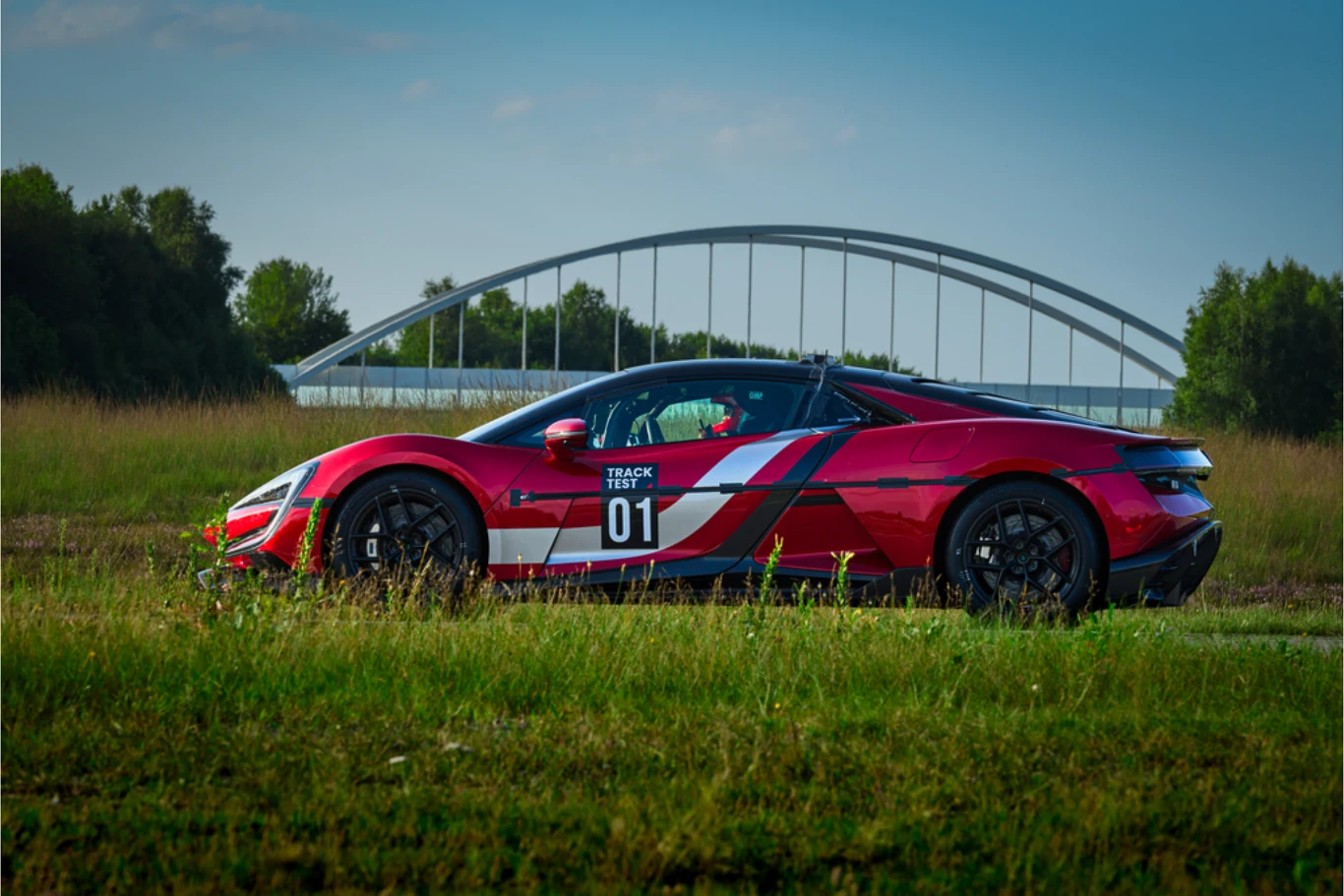 Czerwony samochód sportowy z numerem 01 i oznaczeniami wyścigowymi ustawiony na zielonej trawie, w tle most łukowy oraz drzewa.