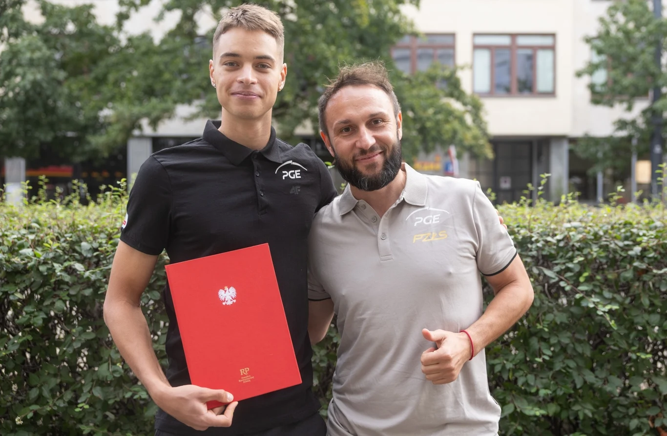 Dwóch mężczyzn stoi razem na tle krzewów i budynku, jeden z nich trzyma czerwone teczkę z godłem Polski, obaj uśmiechnięci, ubrani w koszulki z logo PGE.