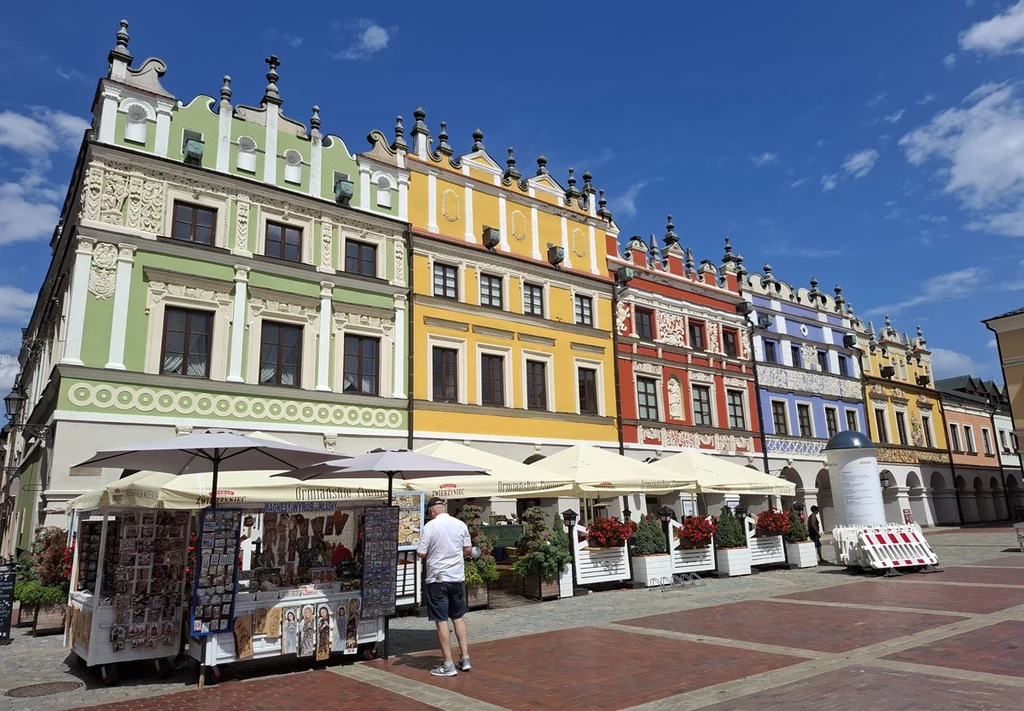 Kolorowe kamienice z bogato zdobionymi fasadami stojące w szeregu przy rynku, przed nimi liczne parasole i stoiska z pamiątkami, pod jednym z nich stoi mężczyzna w jasnych ubraniach, pogodny dzień z błękitnym niebem i chmurami.