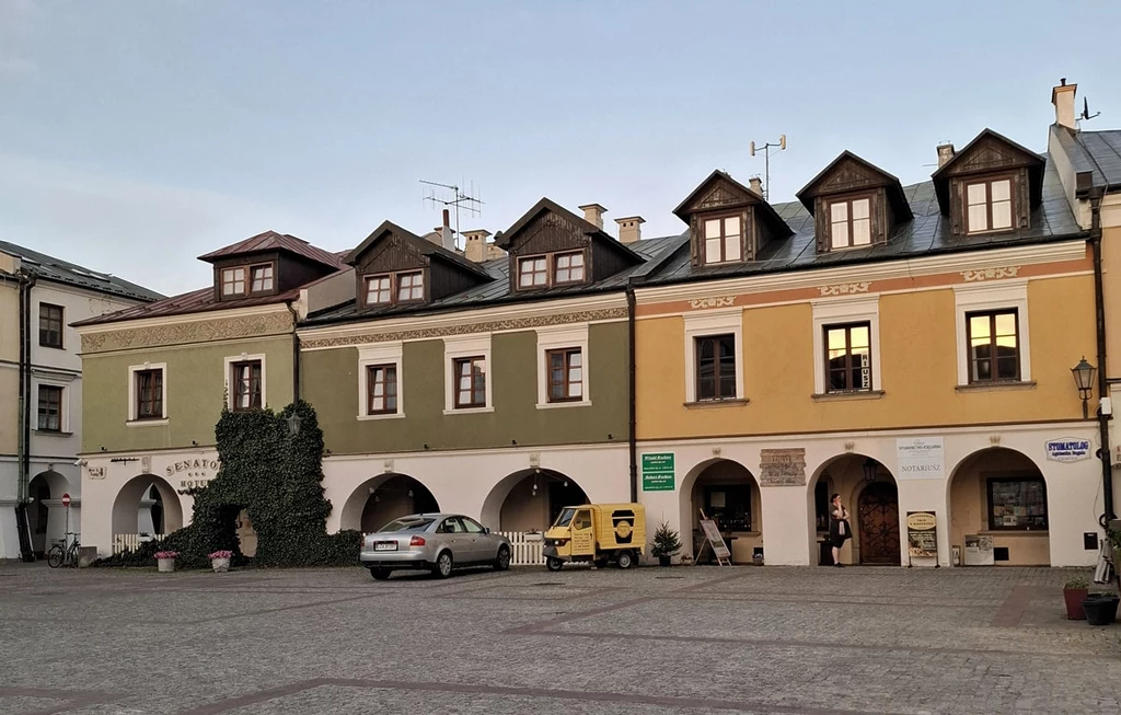 Kolorowe kamienice z arkadami stoją w szeregu przy brukowanym placu, na tle jasnego nieba. Przed budynkami zaparkowany jest samochód osobowy oraz żółty pojazd dostawczy, do jednej z kamienic pnie się gęsty bluszcz.