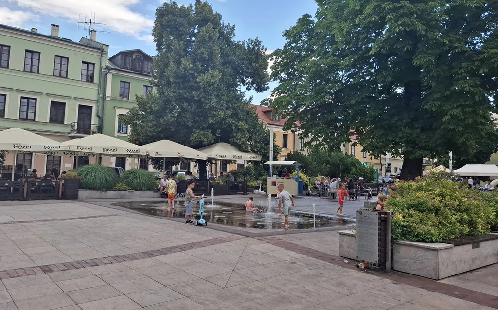 Plac miejski z fontanną, wokół której bawią się dzieci, w tle kamienice, kawiarniane ogródki pod parasolami oraz liczne drzewa zapewniające cień.