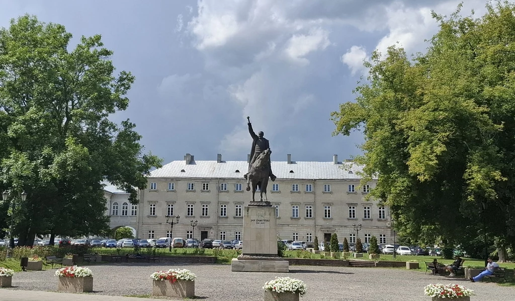 Pomnik jeźdźca na koniu ustawiony na wysokim cokole, w otoczeniu placu z kwiatowymi klombami, drzewa po bokach oraz klasycystyczny budynek w tle z zaparkowanymi samochodami.