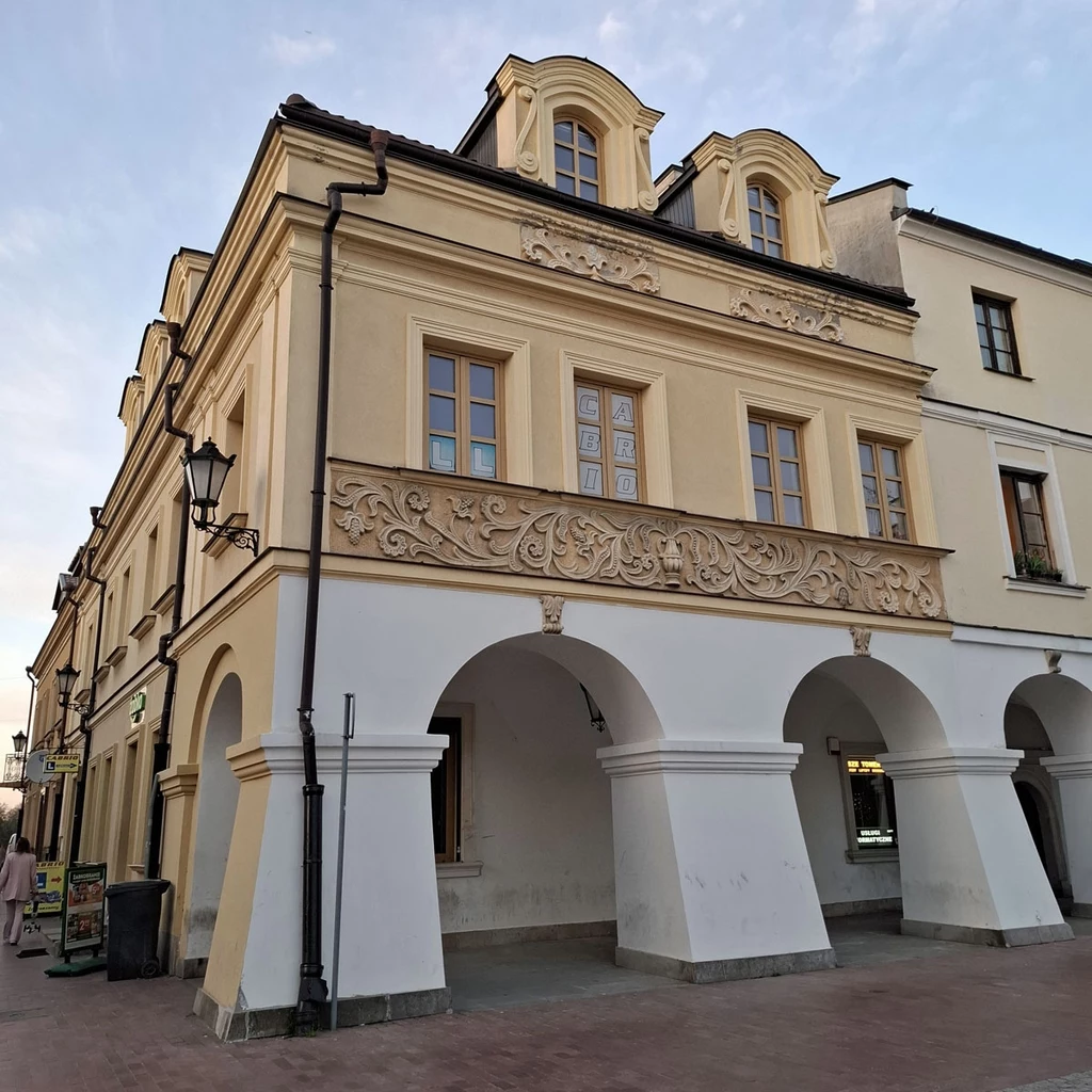 Zabytkowa kamienica z jasną elewacją oraz dekoracyjnymi gzymsami, wykończona łukowymi podcieniami na parterze, zdobiona detalami sztukatorskimi, położona przy brukowanym deptaku.