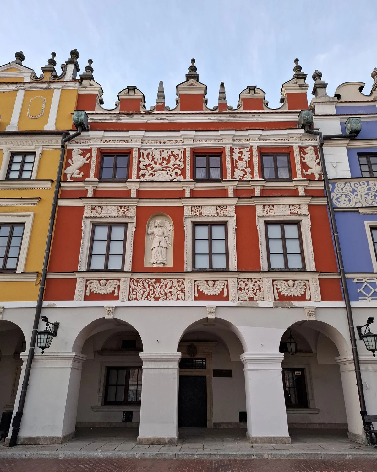 Elewacja zabytkowej kamienicy z czerwonym tynkiem oraz bogatymi dekoracjami sztukatorskimi, zlokalizowana pomiędzy żółtą a niebieską fasadą, z trzema arkadami na parterze i umieszczoną na środku rzeźbą postaci ludzkiej.