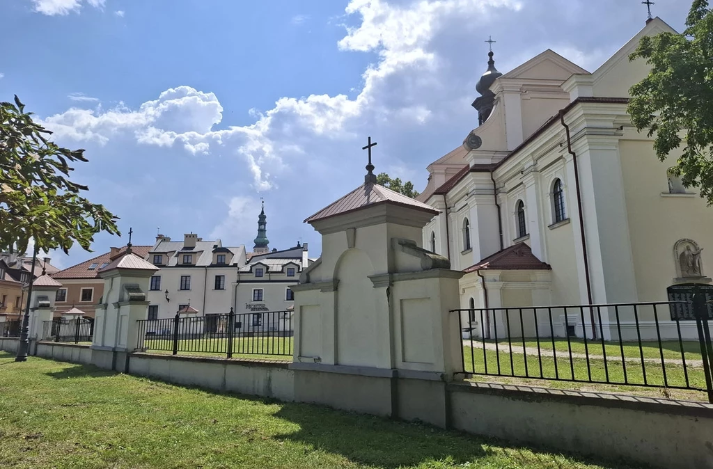 Biała barokowa sylwetka kościoła z charakterystycznymi kopułami i krzyżami, ogrodzona metalowym płotem oraz murkiem, w tle zabudowa mieszkalna i wyraźnie widoczna wieża kościelna, dookoła zieleń i niebo z cumulusami.
