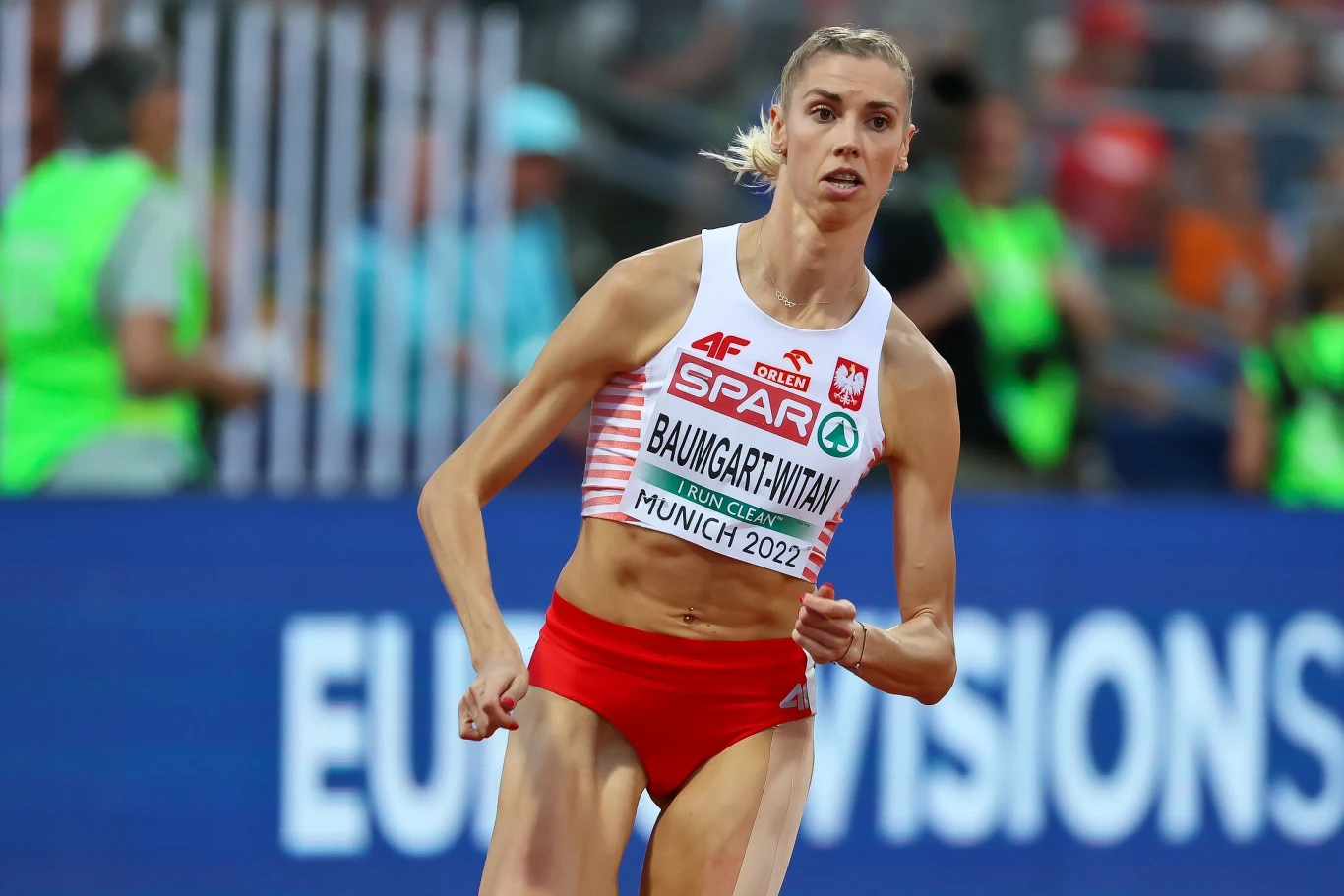 Polska lekkoatletka w czasie biegu na stadionie, ubrana w oficjalny strój reprezentacyjny z widocznym numerem i nazwiskiem, w tle rozmyta publiczność i obsługa wydarzenia.