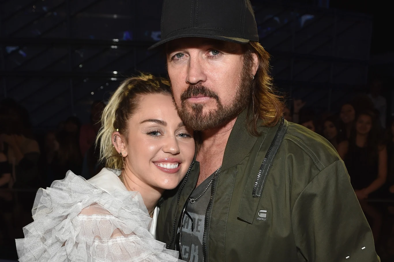 Miley Cyrus ze swoim ojcem, Billym Rayem Kobieta w białej, ozdobnej bluzce przytula się do mężczyzny w zielonej kurtce i czapce, oboje uśmiechnięci, pozują razem do zdjęcia, w tle niewyraźne postaci.