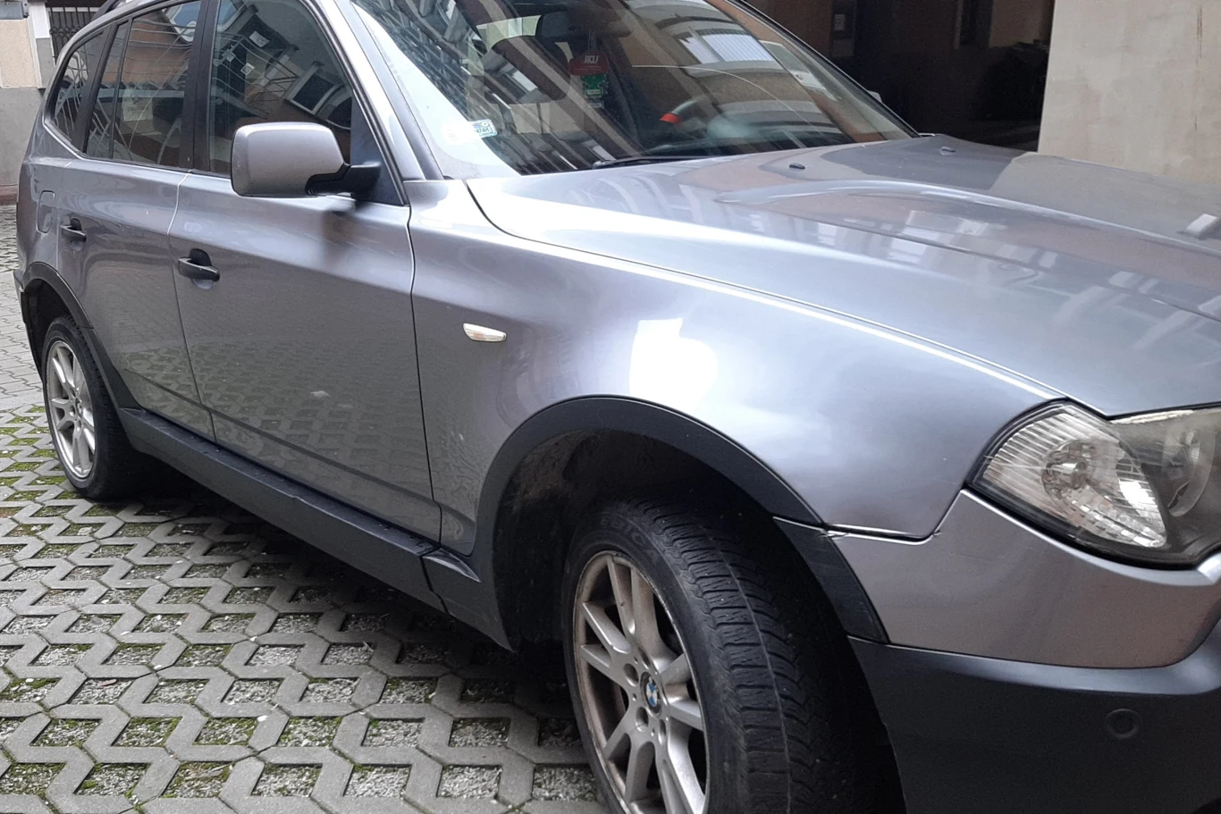 BMW X3 z 2005 roku za 5 tys. zł. Fot. US w Tarnowie Srebrny samochód osobowy typu SUV zaparkowany na kostce brukowej między budynkami, refleksy światła odbijają się na karoserii pojazdu.