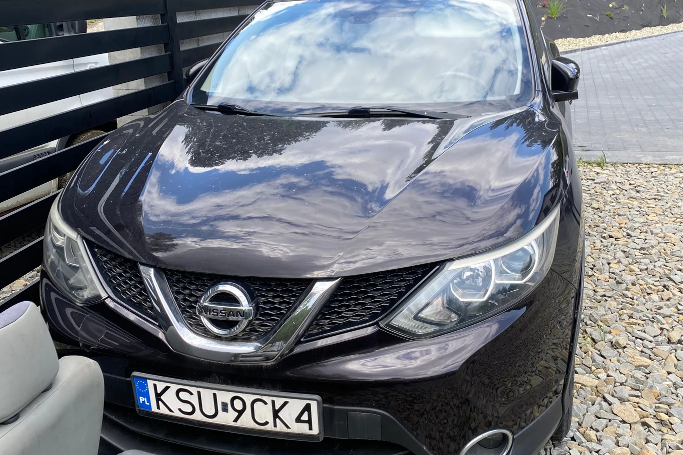 Nissan Qashqai z 2014 roku. Jego cena wywoławcza to 22 500 zł. | Fot. US w Suchej Beskidzkiej Czarny samochód marki Nissan ustawiony na podjeździe wyłożonym kamieniami, obok czarnego płotu, z widoczną polską tablicą rejestracyjną z literami KSU.