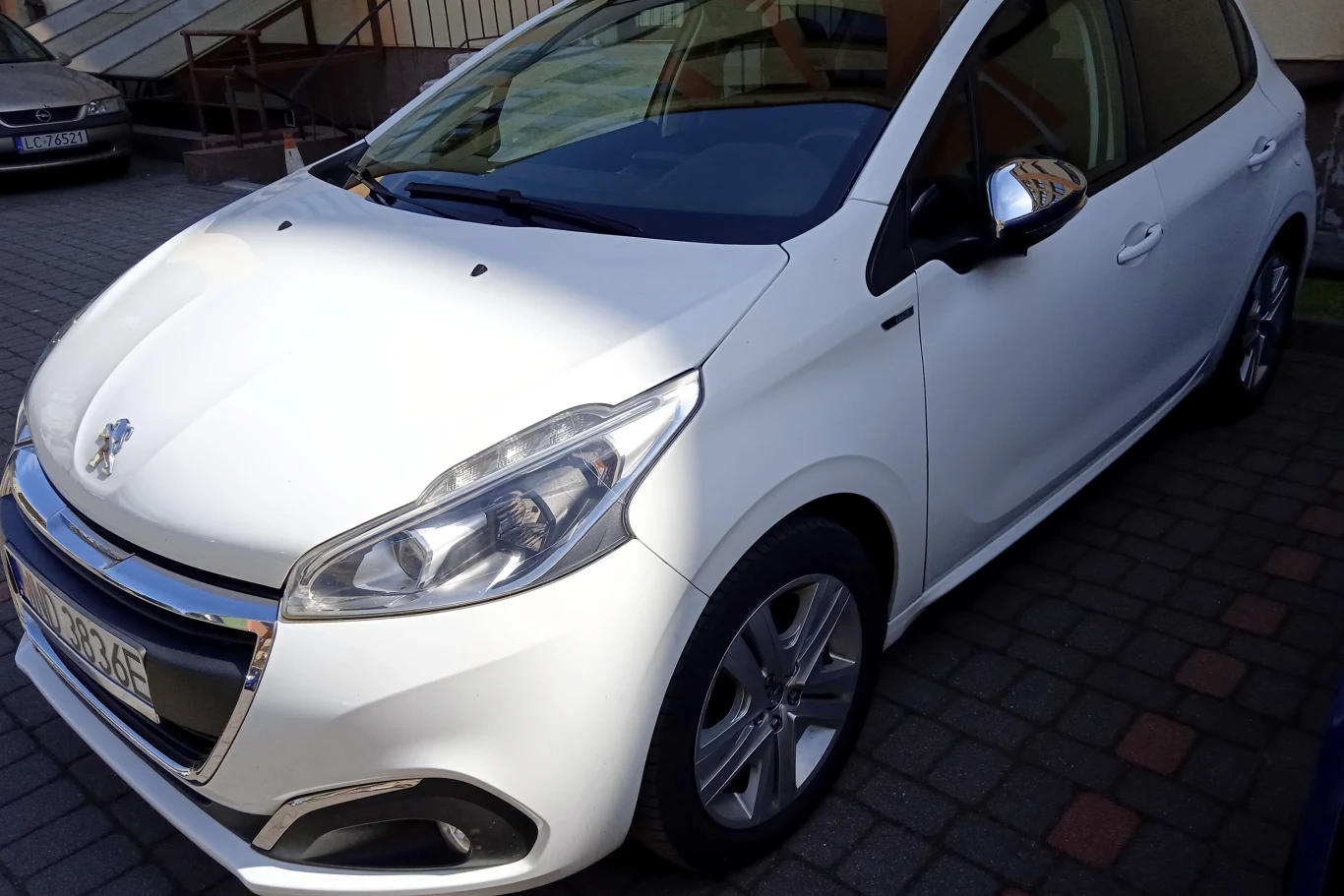 Peugeot 208 z 2016 roku w cenie wywoławczej 17 025 zł. | Fot. US w Nowym Dworze Mazowieckiem Biały samochód osobowy marki Peugeot 208 zaparkowany na brukowanym parkingu obok budynku, widoczna przednia i boczna część pojazdu.