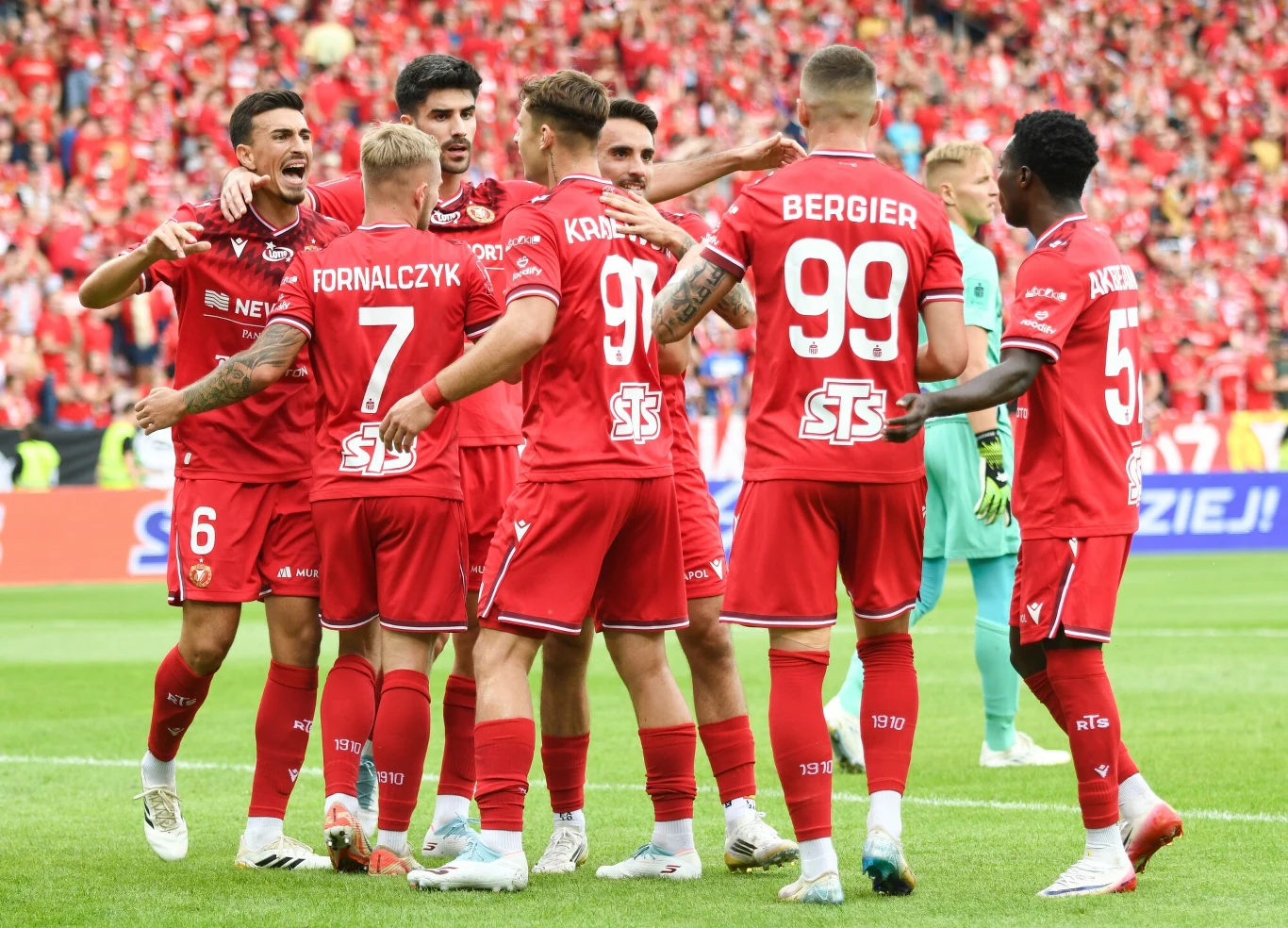 Grupa piłkarzy w czerwonych strojach celebruje zdobycie bramki na boisku, otoczona rozemocjonowanymi kibicami na trybunach.