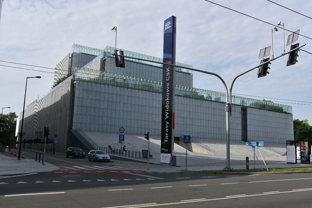 Nowoczesny budynek główny Centrum Nauki Kopernik pokryty szklanymi panelami, położony przy skrzyżowaniu z widocznymi sygnalizatorami świetlnymi i szeroką ulicą, otoczony zielenią i elementami miejskiej infrastruktury.