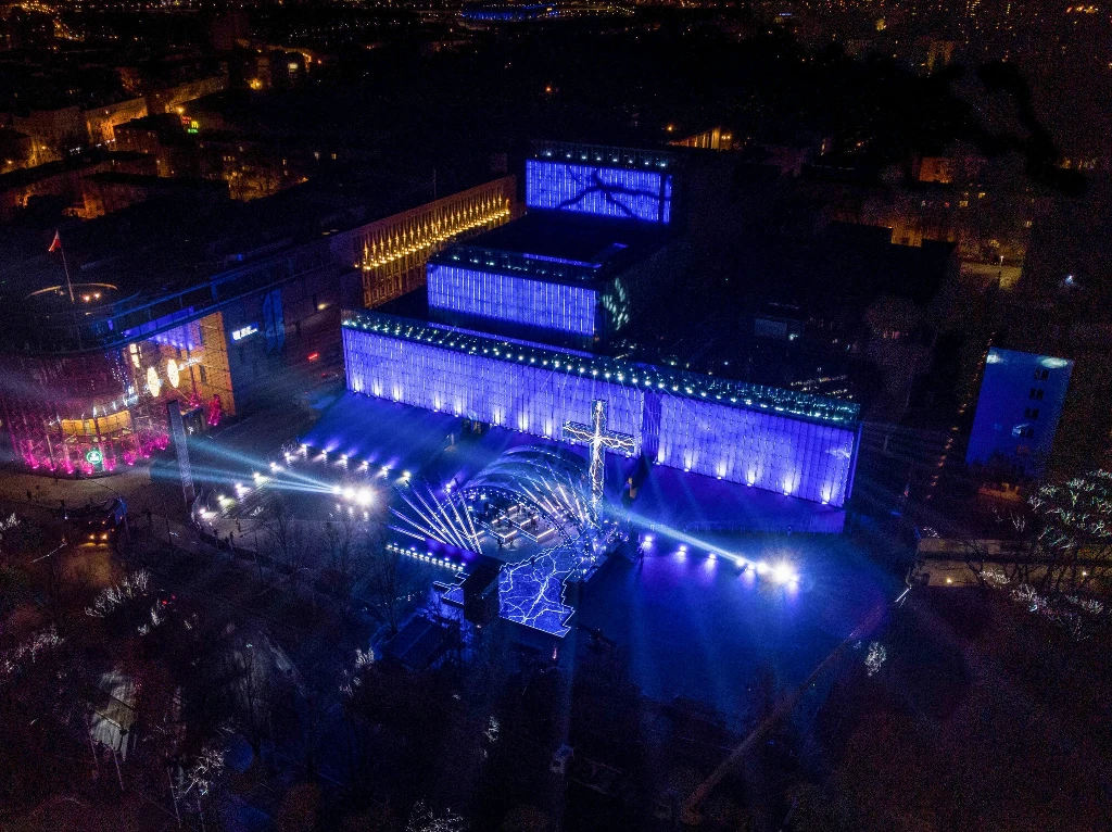 Nocna panorama centrum miasta z podświetlonym na niebiesko dużym budynkiem, przed którym znajduje się scena koncertowa oświetlona wieloma reflektorami, na placu zgromadzeni ludzie.