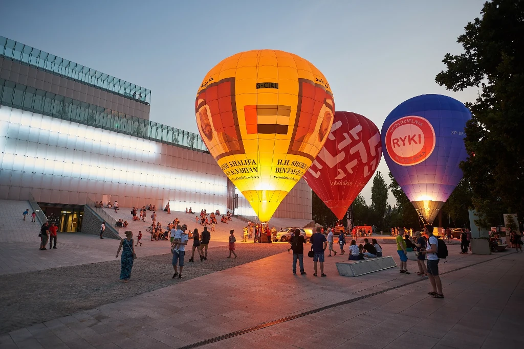 Kolorowe balony na gorące powietrze unoszą się naprzeciwko nowoczesnego budynku, wokół nich zgromadzeni są ludzie obserwujący wydarzenie i spacerujący po placu, pod wieczornym niebem.