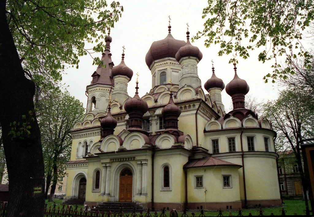 Cerkiew z licznymi cebulastymi kopułami o jasnożółtych ścianach i brązowych dachach, otoczona zielonymi drzewami i metalowym ogrodzeniem, z drewnianymi drzwiami wejściowymi na centralnej fasadzie.