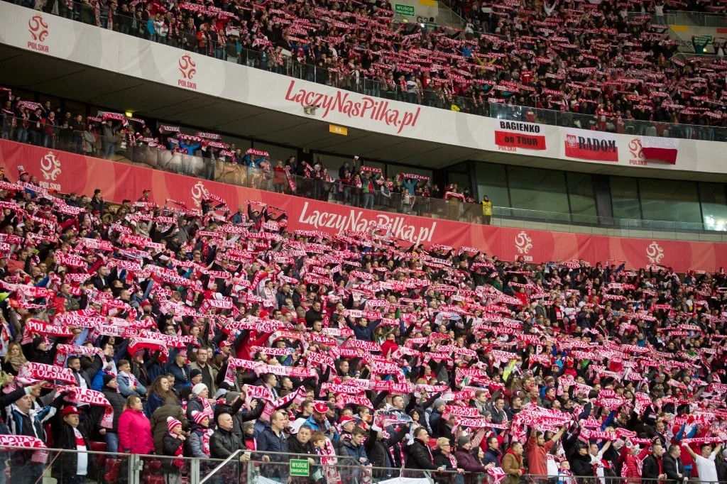 Tłum kibiców na stadionie ubranego w czerwono-białe barwy, trzymających nad głowami szaliki z napisem Polska, atmosfera wspólnego wsparcia i entuzjazmu sportowego.