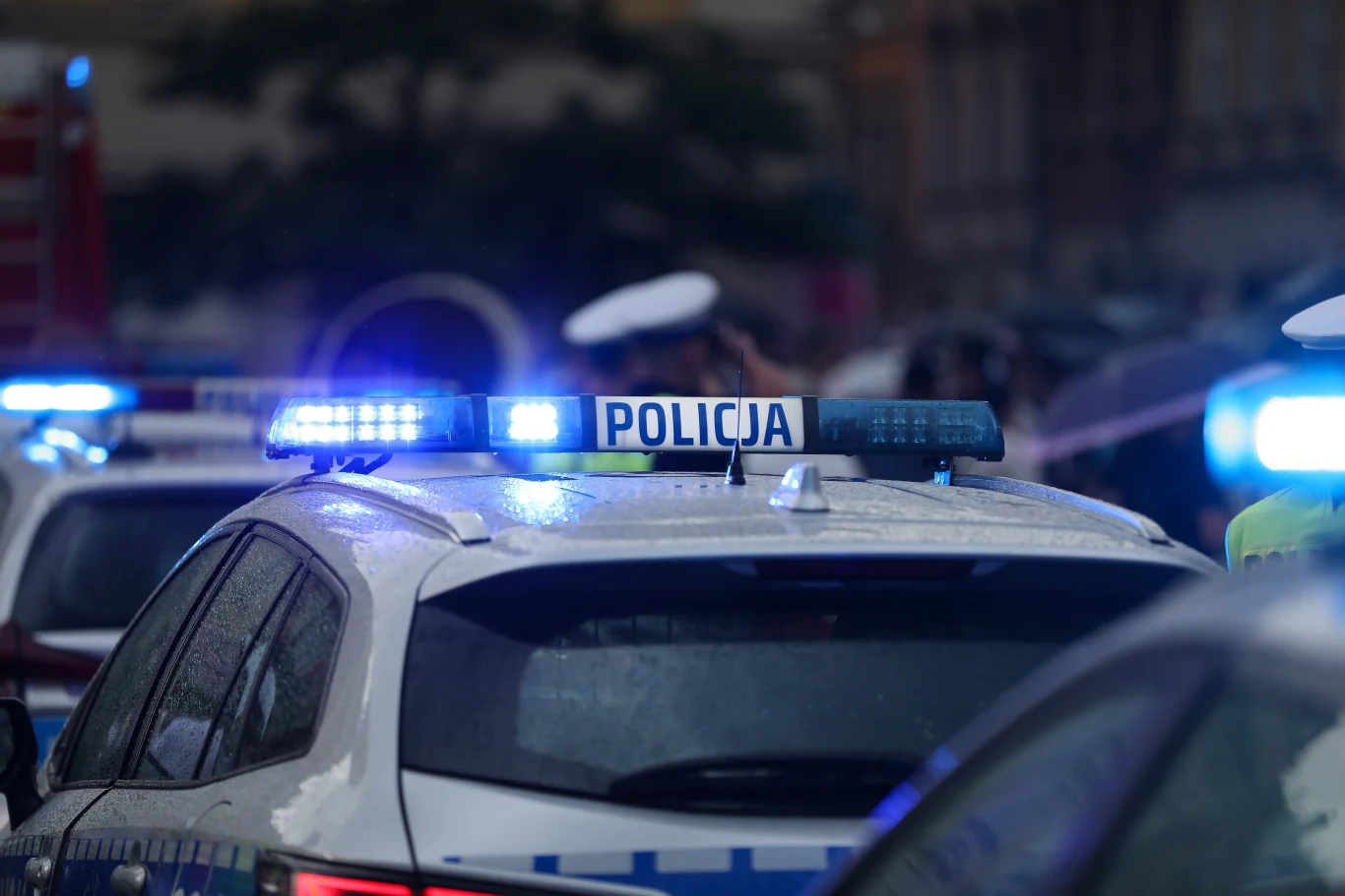 Polityk z Lęborka spowodował kolizję pod wpływem alkoholu Radiowóz policyjny z włączonymi niebieskimi światłami ostrzegawczymi, funkcjonariusze w białych czapkach na tle zatłoczonej ulicy miejskiej.