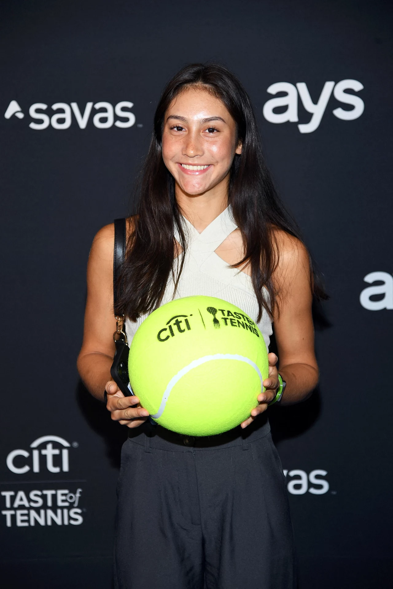 Valerie Glozman Młoda kobieta z długimi ciemnymi włosami trzyma dużą, neonowo-żółtą piłkę tenisową z logotypem Citi Taste of Tennis, stojąc na tle ściany z logami sponsorów.