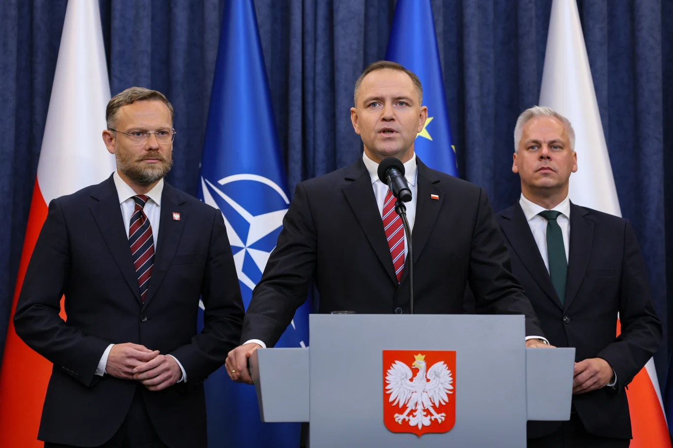 Karol Nawrocki może zawetować podwyżkę akcyzy, planowaną przez rząd Konferencja prasowa z udziałem trzech mężczyzn w garniturach stojących przed mównicą z godłem Polski, w tle widoczne flagi Polski, NATO oraz Unii Europejskiej. Centralnie ustawiony mężczyzna przemawia do mikrofonu.