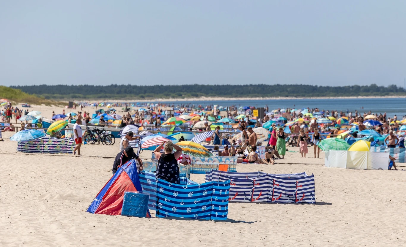 Ciało znalezione na plaży w Jastarni należy do poszukiwanego 18-latka (zdjęcie ilustracyjne) Tłum ludzi odpoczywających na piaszczystej plaży, kolorowe parawany i parasole rozstawione nad brzegiem morza, słoneczna pogoda oraz spokojna linia horyzontu.