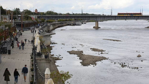 bulwary nadrzeczne z licznymi spacerowiczami, rzeka o obniżonym stanie wody odsłaniająca kamieniste brzegi, most z przejeżdżającym pociągiem, po lewej stronie alejka ze stolikami i roślinnością