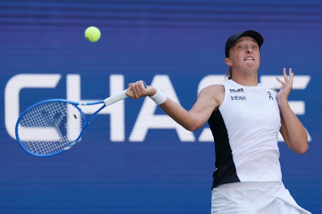 Iga Świątek w 2. rundzie US Open Tenisistka uderzająca piłkę podczas meczu tenisowego na tle niebieskiego baneru z widoczną częścią logo sponsora.