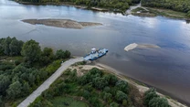 Przeprawa promowa przez szeroką rzekę z wyłaniającymi się piaszczystymi łachami, otoczona gęstą zielenią i lasami, niewielka droga prowadzi do brzegu, nad całością rozciąga się spokojny krajobraz przyrodniczy.