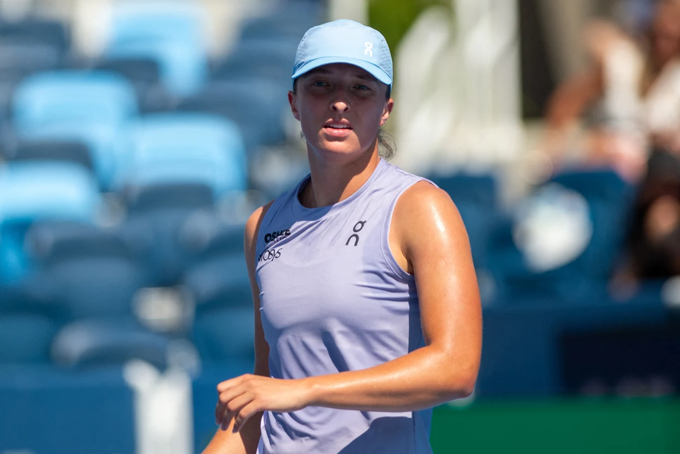 Kobieta w sportowym stroju i niebieskiej czapce z daszkiem podczas meczu tenisowego, na tle rozmazanych trybun w słoneczny dzień.