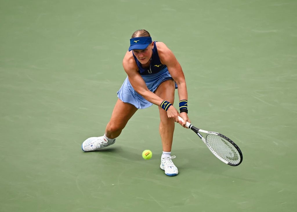 Suzan Lamens Kobieta w sportowym stroju, skupiona na odbiciu nadlatującej piłki tenisowej rakietą na korcie o zielonej nawierzchni.