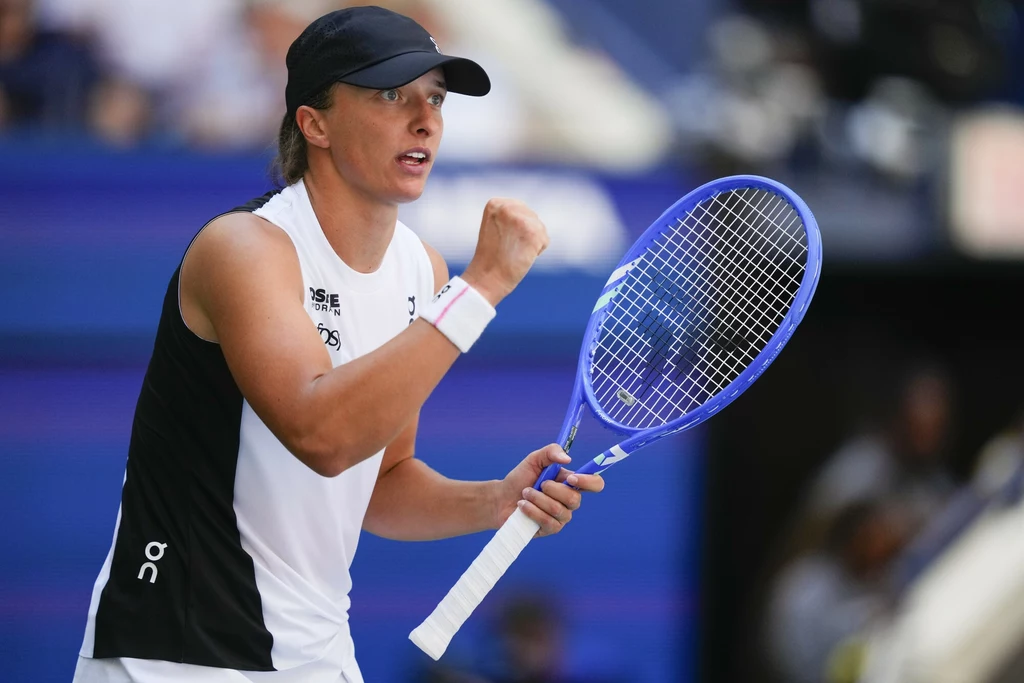 Tenisistka w sportowym stroju i czapce, z uniesioną zaciśniętą pięścią, trzyma niebieską rakietę tenisową, na jej twarzy widać determinację, a w tle rozmyte trybuny stadionu.