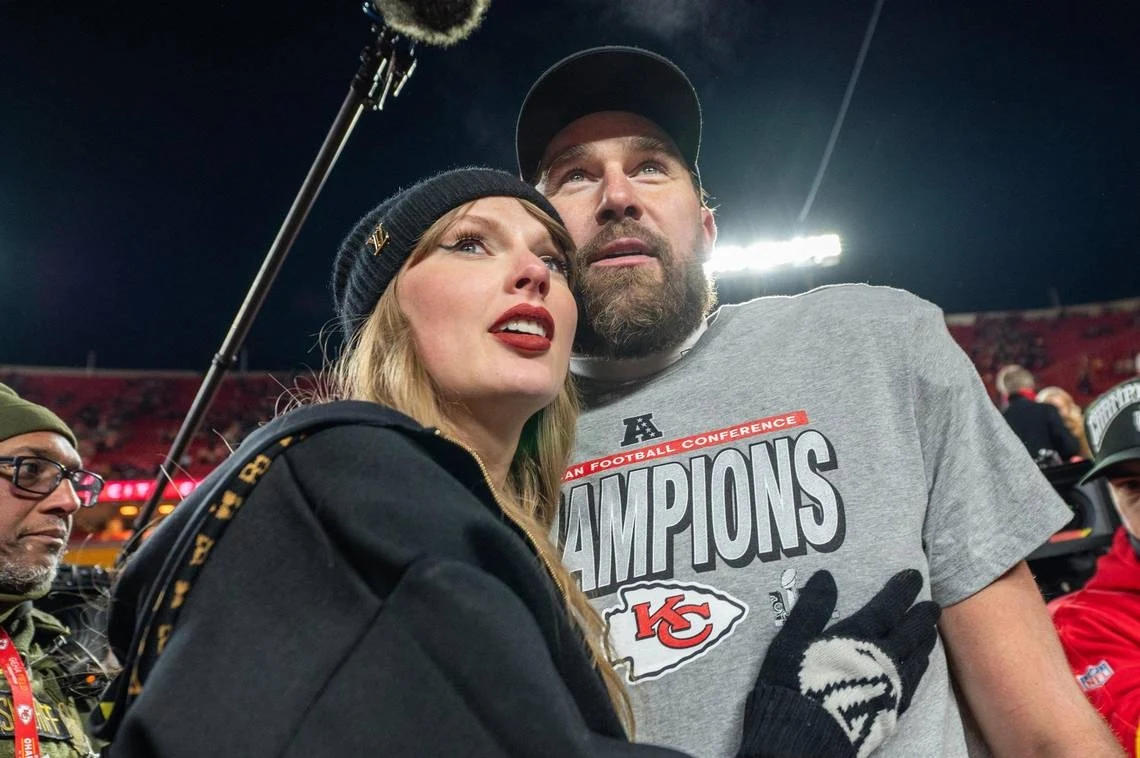 Taylor Swift zaręczyła się z Travisem Kelce Młoda kobieta ubrana w czarną czapkę oraz mężczyzna w sportowej bluzie z napisem 'Champions' i logo Kansas City Chiefs pozują razem na stadionie, w tle widać światła oraz innych ludzi.