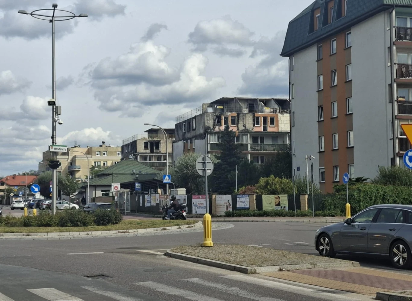 Uszkodzony budynek mieszkalny z częściowo zniszczonym dachem oraz wypaloną elewacją, pośród innych, mniej uszkodzonych budynków w miejskim krajobrazie, na skrzyżowaniu z kilkoma samochodami i znakami drogowymi.