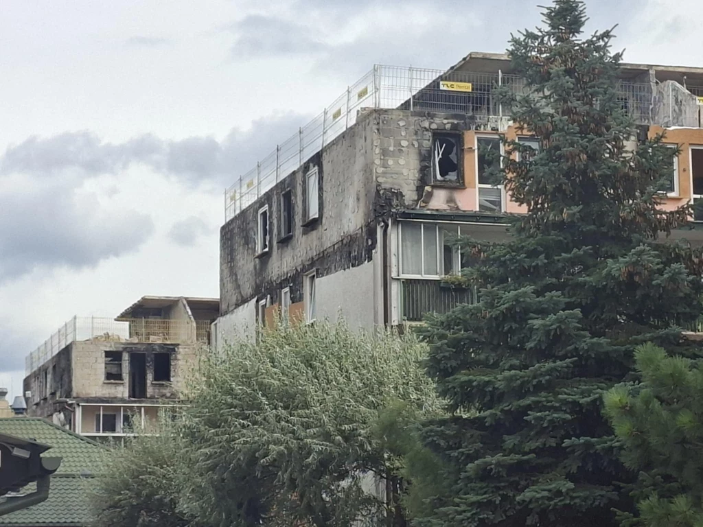 Tak pod koniec sierpnia wygląda blok, który spłonął w Ząbkach Spalony i zniszczony wielorodzinny budynek mieszkalny z widocznymi śladami ognia, uszkodzeniami elewacji i powybijanymi szybami, na pierwszym planie częściowo zasłonięty przez drzewo.