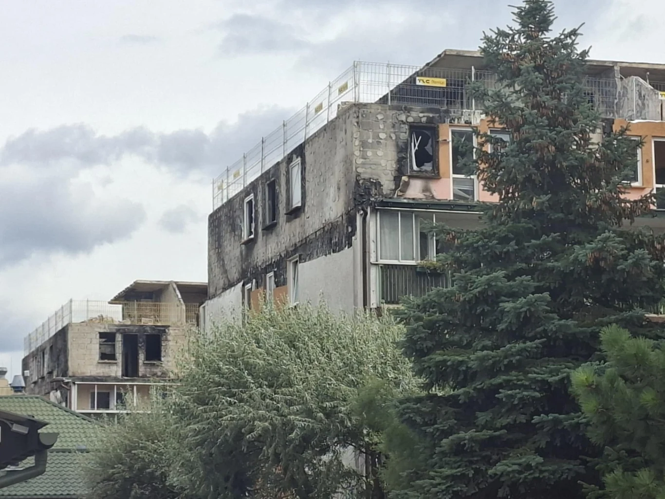Spalony i zniszczony wielorodzinny budynek mieszkalny z widocznymi śladami ognia, uszkodzeniami elewacji i powybijanymi szybami, na pierwszym planie częściowo zasłonięty przez drzewo.