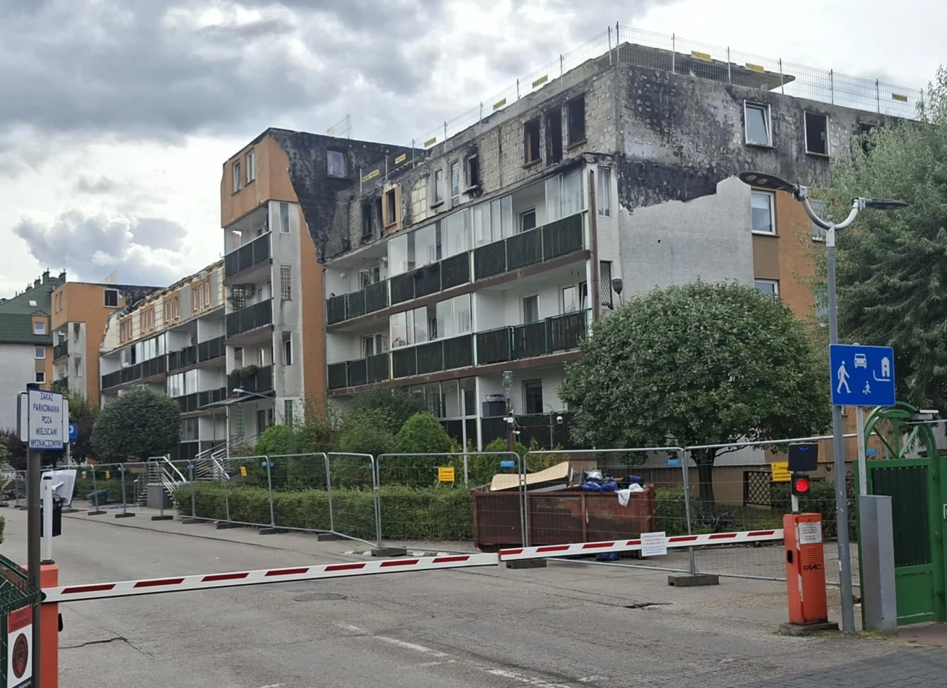 Uszkodzony po pożarze blok mieszkalny z osmolonymi ścianami górnych pięter, otoczony płotem zabezpieczającym, w tle chmury, widoczna szlabanowa brama wjazdowa oraz oznakowanie dla pieszych.
