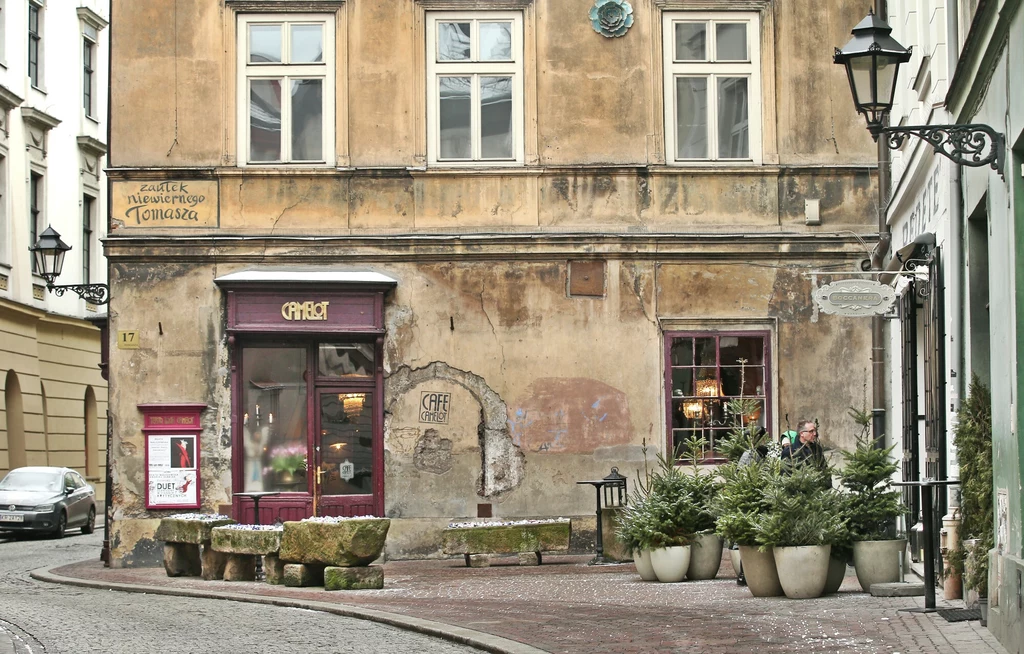 Zaułek Niewiernego Tomasza w Krakowie Stara, kamienista uliczka z zabytkową kamienicą o zniszczonej elewacji, kawiarnią z czerwonym wejściem oraz grupą donic z iglakami ustawioną przy ścianie budynku. Dwie osoby siedzą za oknem w kawiarni.