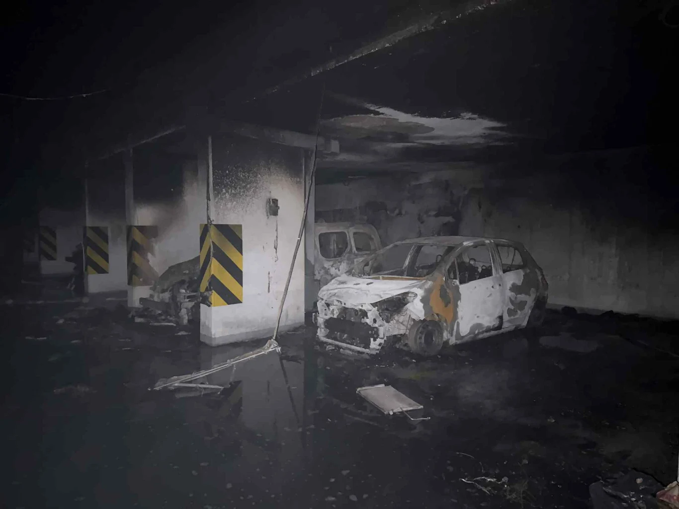 Spalone samochody w podziemnym garażu, osmolone ściany, zniszczone elementy infrastruktury, wszechobecny dym i czarne zacieki na suficie.