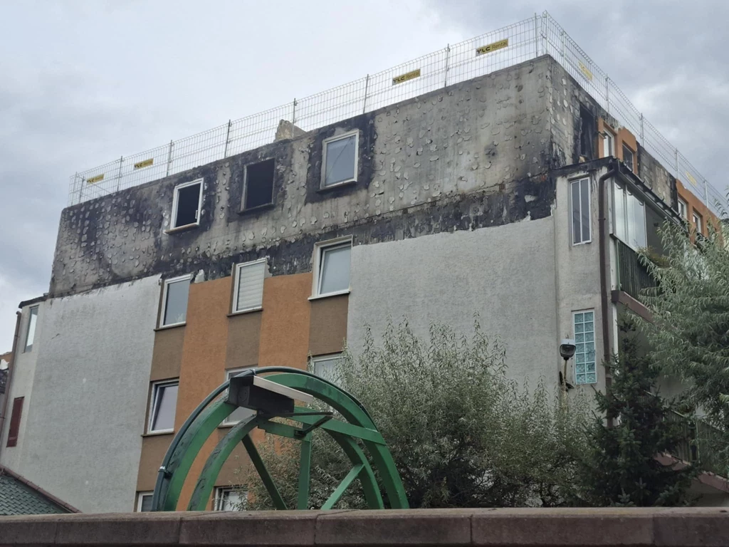 Nie wiadomo, czy blok w Ząbkach będzie nadawał się jeszcze do zamieszkania Uszkodzona elewacja budynku mieszkalnego z widocznymi śladami pożaru na ostatnim piętrze. Na dachu zamontowano tymczasowe ogrodzenie zabezpieczające. Na pierwszym planie znajduje się fragment zielonej konstrukcji, częściowo zasłoniętej przez roślinność.