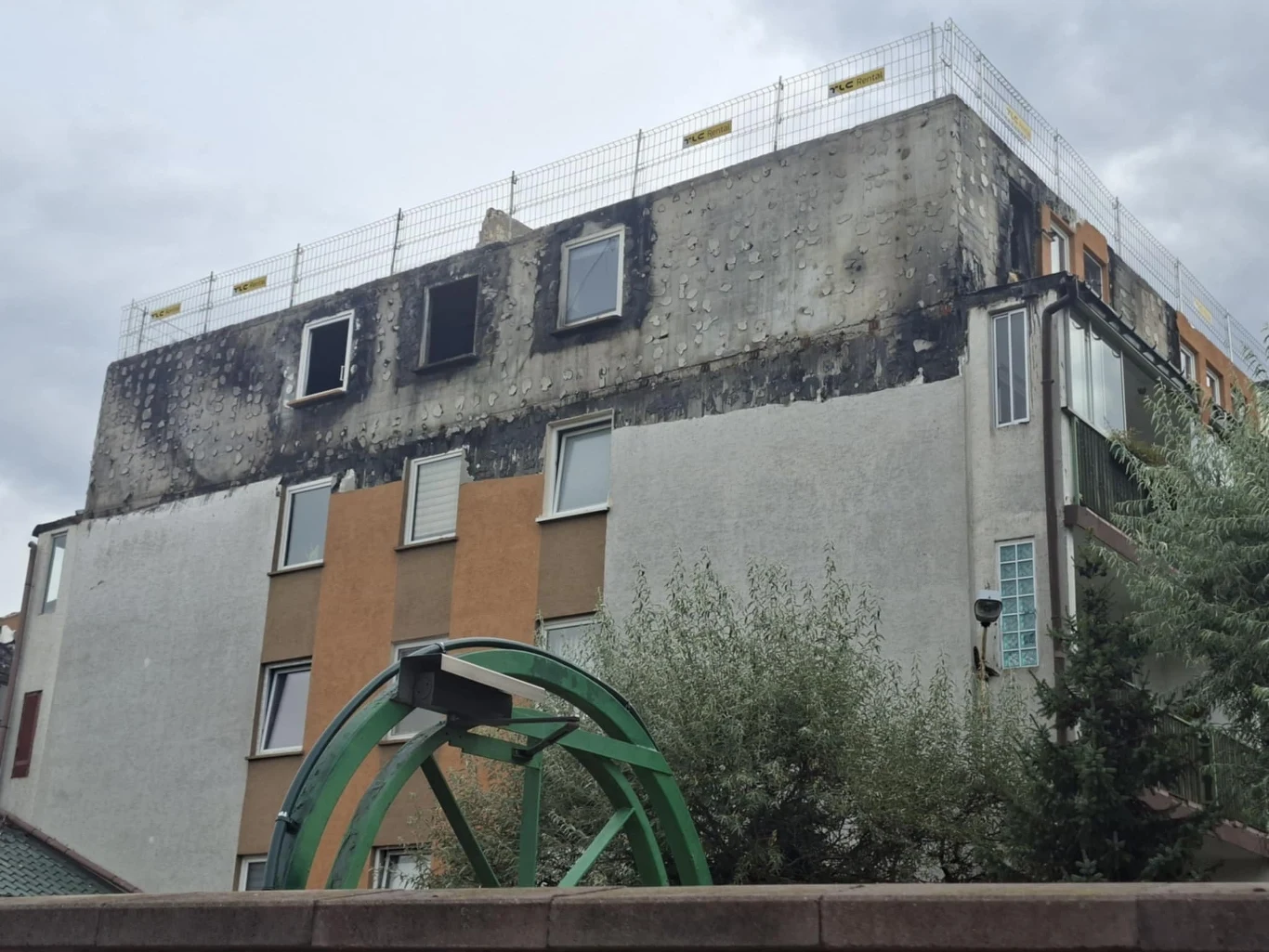 Uszkodzona elewacja budynku mieszkalnego z widocznymi śladami pożaru na ostatnim piętrze. Na dachu zamontowano tymczasowe ogrodzenie zabezpieczające. Na pierwszym planie znajduje się fragment zielonej konstrukcji, częściowo zasłoniętej przez roślinność.