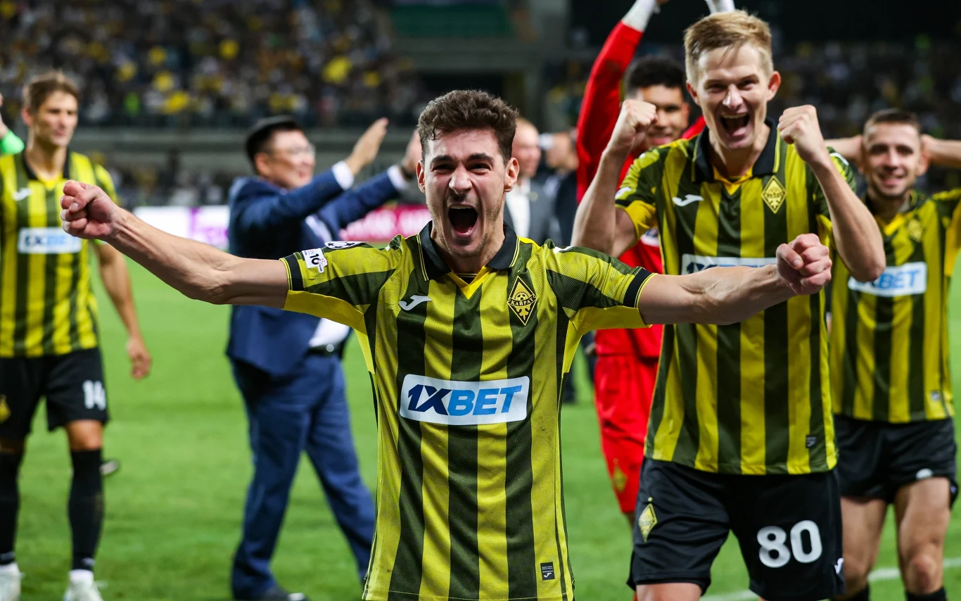 Kairat Ałmaty w Lidze Mistrzów Radość grupy piłkarzy w żółto-czarnych koszulkach z herbem klubu po zwycięstwie, z szerokimi uśmiechami i gestami triumfu, tłum w tle oraz obecność trenerów i sztabu na boisku.