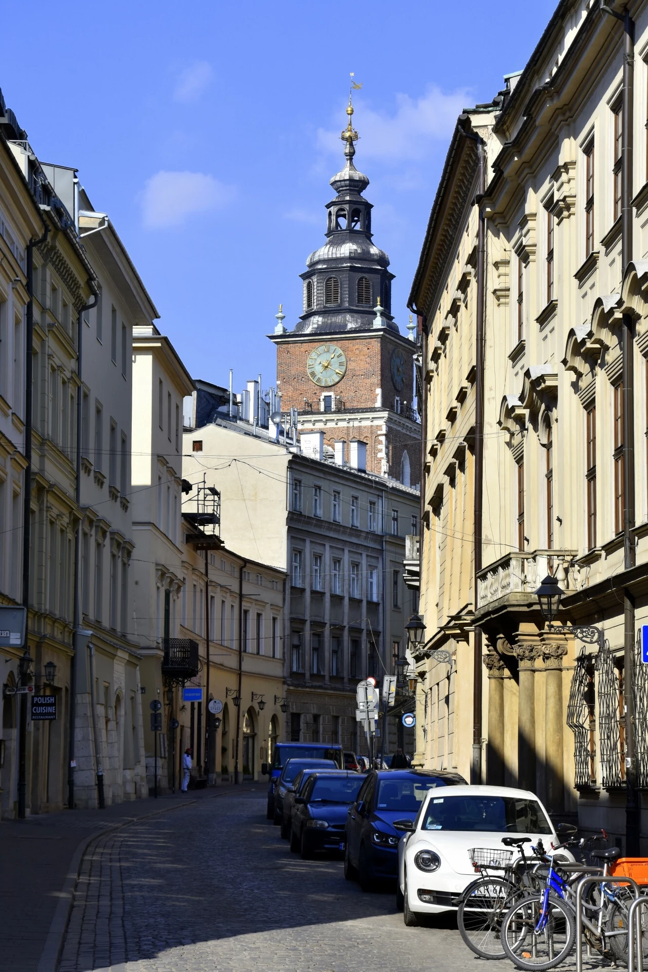 Ul. Bracka w Krakowie Wąska brukowana ulica w otoczeniu historycznych kamienic, w tle widoczna wieża ratusza z zegarem, na ulicy parkują samochody i stoją rowery, słoneczna pogoda podkreśla detale architektury.
