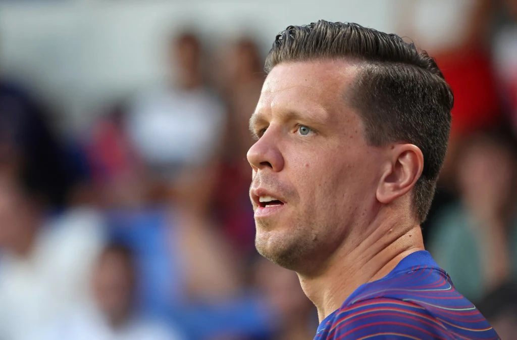 Wojciech Szczęsny Mężczyzna w sportowej koszulce w niebiesko-czerwone paski, stoi w profilu, w tle rozmyty tłum ludzi na trybunach.