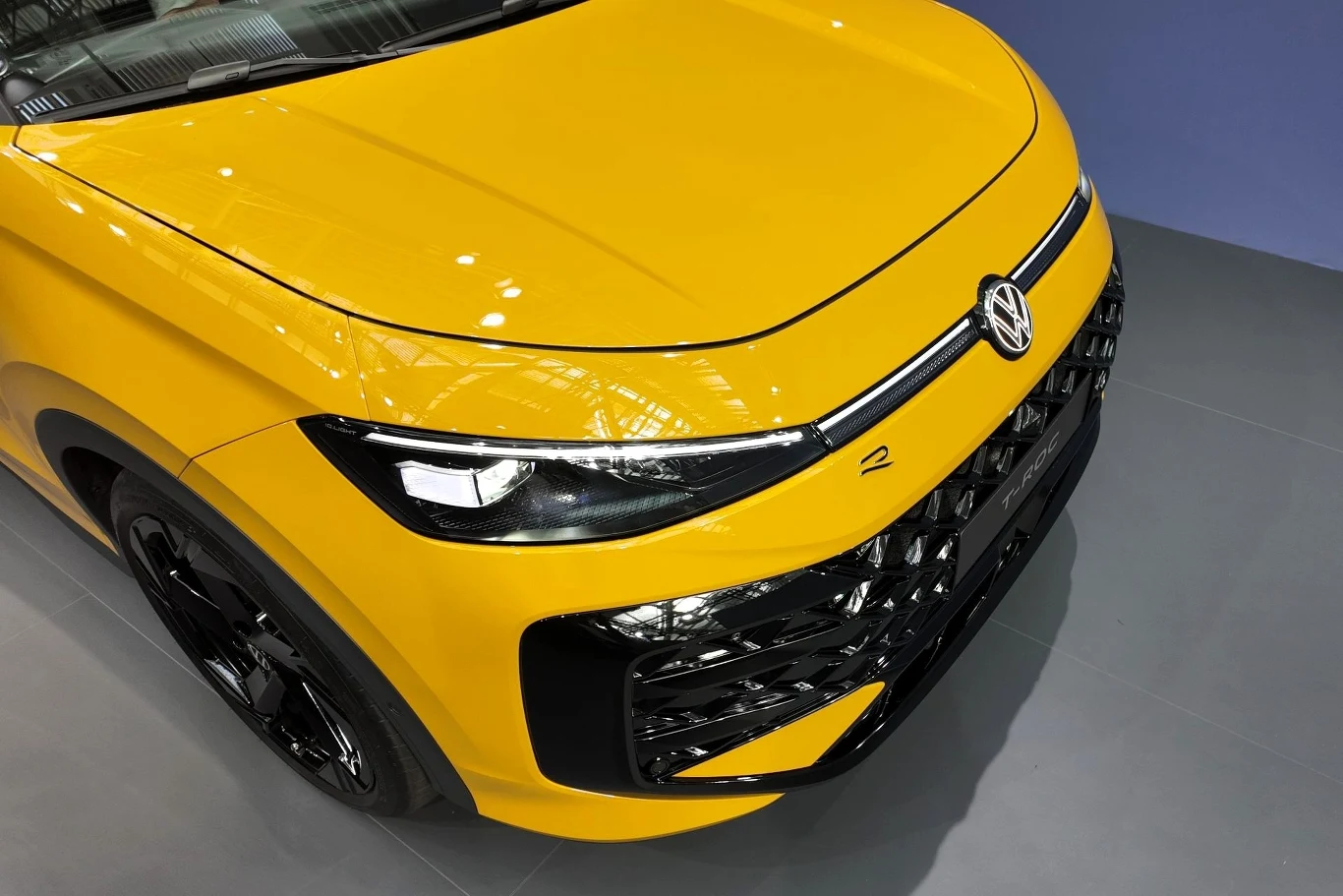 Przód żółtego samochodu Volkswagen z nowoczesnym oświetleniem LED i lśniącym wykończeniem, widoczny fragment maski, zderzaka oraz reflektorów, całość na tle neutralnej, szarej podłogi.