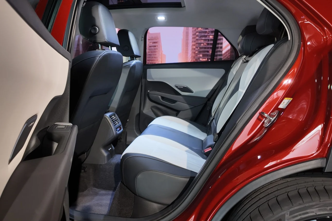 Tylny rząd siedzeń w nowoczesnym samochodzie osobowym, tapicerka w odcieniach szarości i bieli, szeroko otwarte drzwi, widoczne miejskie budynki za oknem.