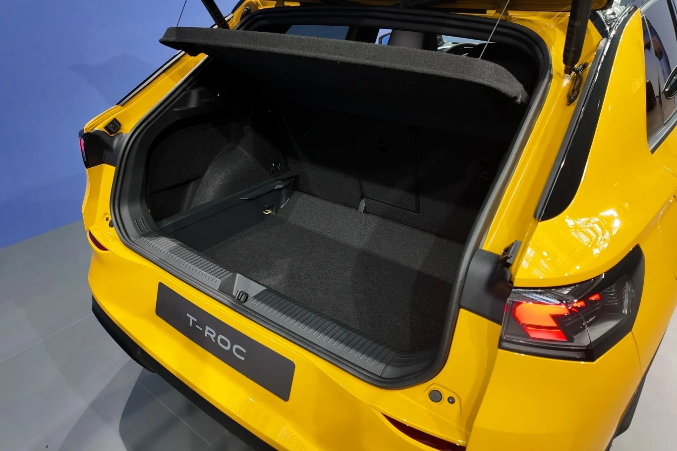 Otwarty bagażnik żółtego samochodu Volkswagen T-Roc z widocznym dużym przestronnym wnętrzem oraz podniesioną półką bagażową, światła tylne i fragment tylnej klapy samochodu.