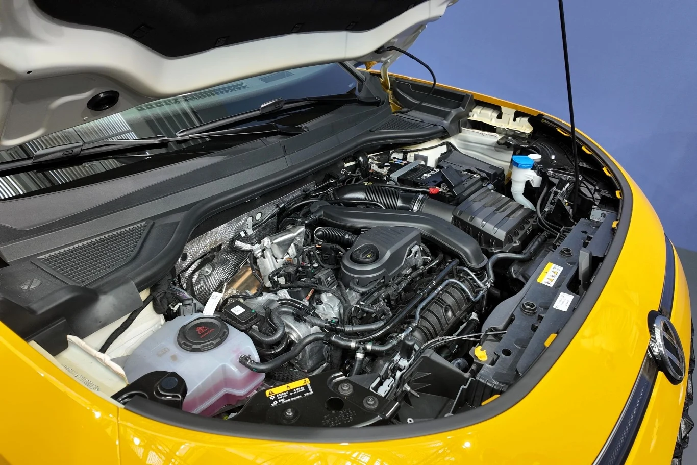 Otwarta maska żółtego samochodu, pod którą widoczny jest nowoczesny silnik spalinowy z wyraźnie zaznaczonymi przewodami, zbiornikami płynów oraz plastikową osłoną silnika.