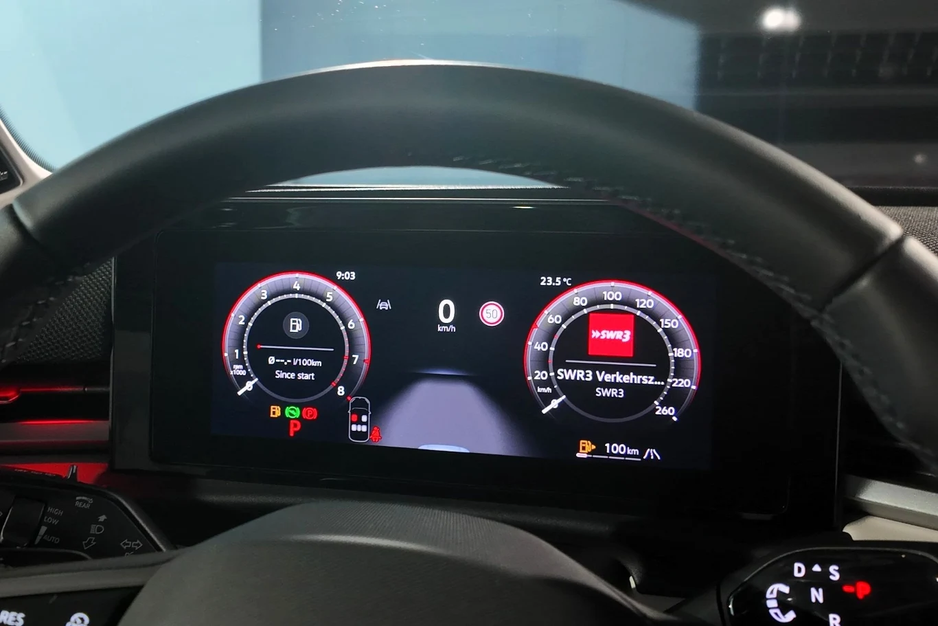 Cyfrowy wyświetlacz deski rozdzielczej samochodu prezentujący liczniki prędkości, obrotomierz, wskaźnik paliwa oraz system nawigacji i informacje radiowe, wokół kierownica w centralnej części kadru.