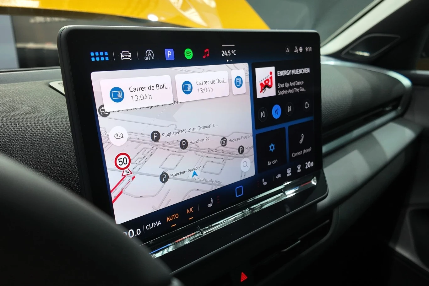 Cyfrowy ekran dotykowy w samochodzie z wyświetloną mapą nawigacji, informacjami o trasie i aktualną temperaturą. Po prawej stronie widać również interfejs do obsługi radia i klimatyzacji.