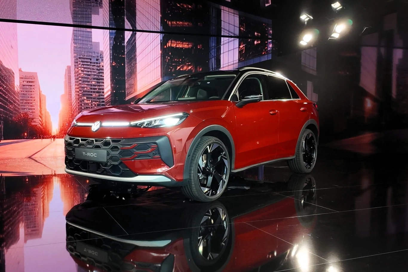 Czerwony samochód osobowy typu SUV marki Volkswagen stoi na błyszczącej, czarnej scenie, w tle wyświetlane są wysokie nowoczesne budynki oraz światła studyjne, które podkreślają sylwetkę pojazdu.